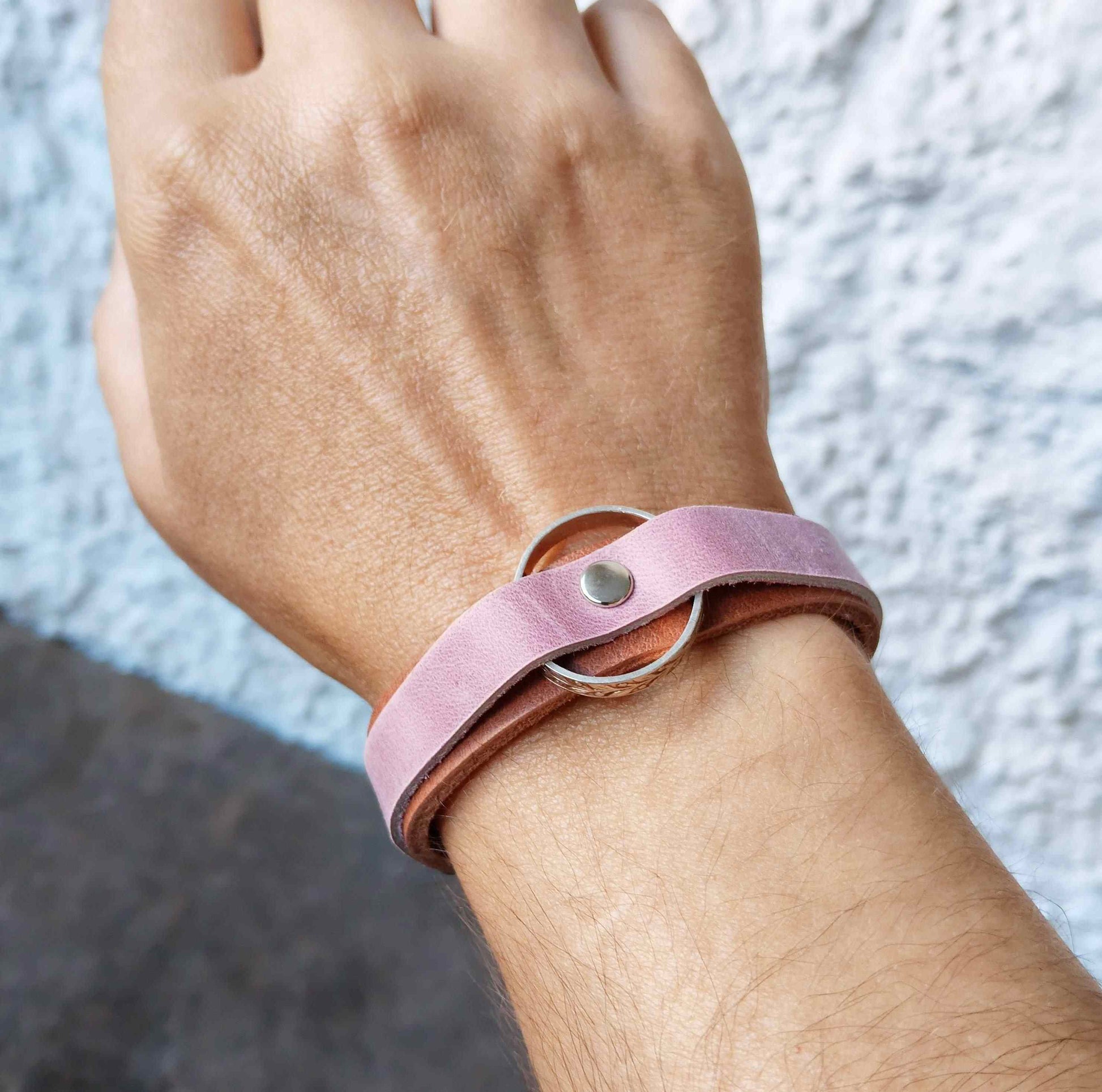 A close-up of a person's wrist wearing a double-band leather bracelet. The top band is a dusty pink suede-like material with a silver metal ring and snap closure, while the bottom band is a warm brown leather.