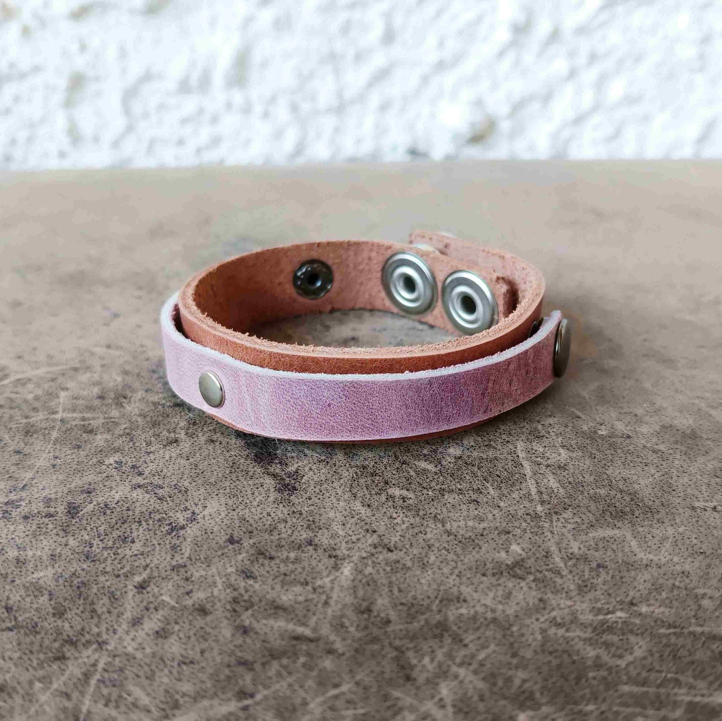A handmade leather wrap bracelet in a dusty pink and light brown color, featuring silver-tone metal snaps and rivets, resting on a textured gray surface.