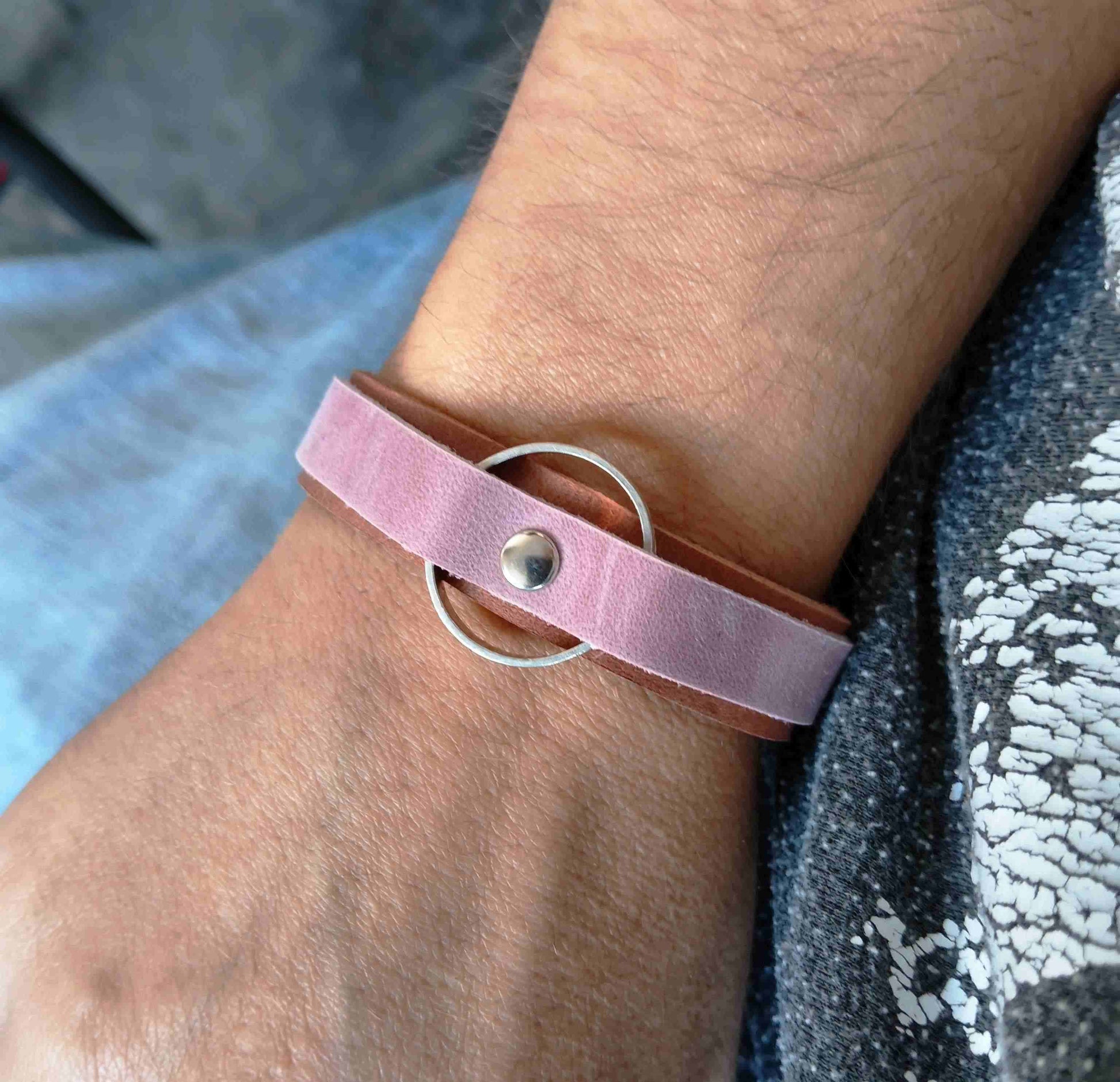 A close-up of a person's wrist wearing a pink suede-like wrap bracelet with a silver metal O-ring and a small silver rivet.