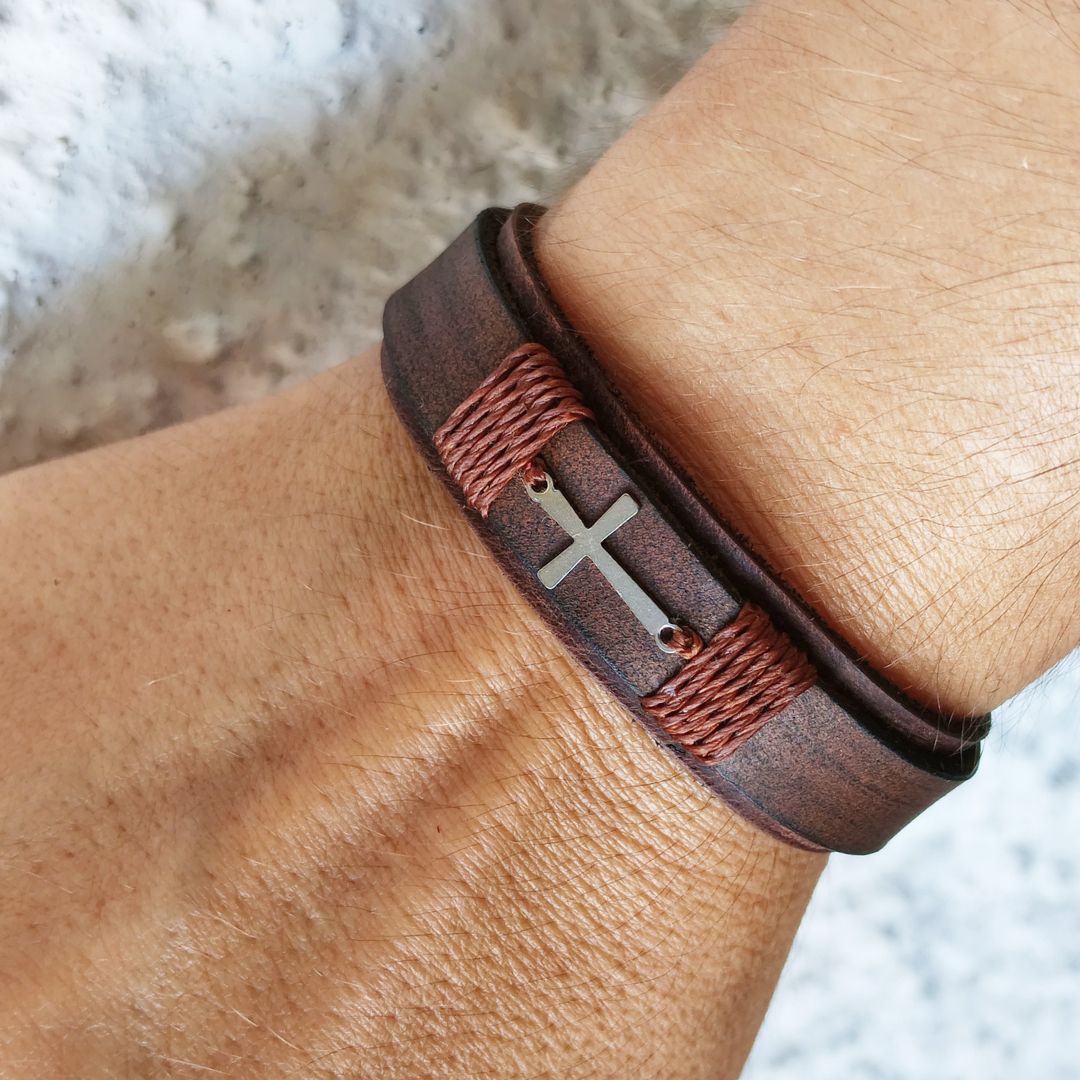 A close-up of a dark brown leather wrap bracelet worn on a wrist, featuring a silver-toned cross charm and textured brown cord wrapping.