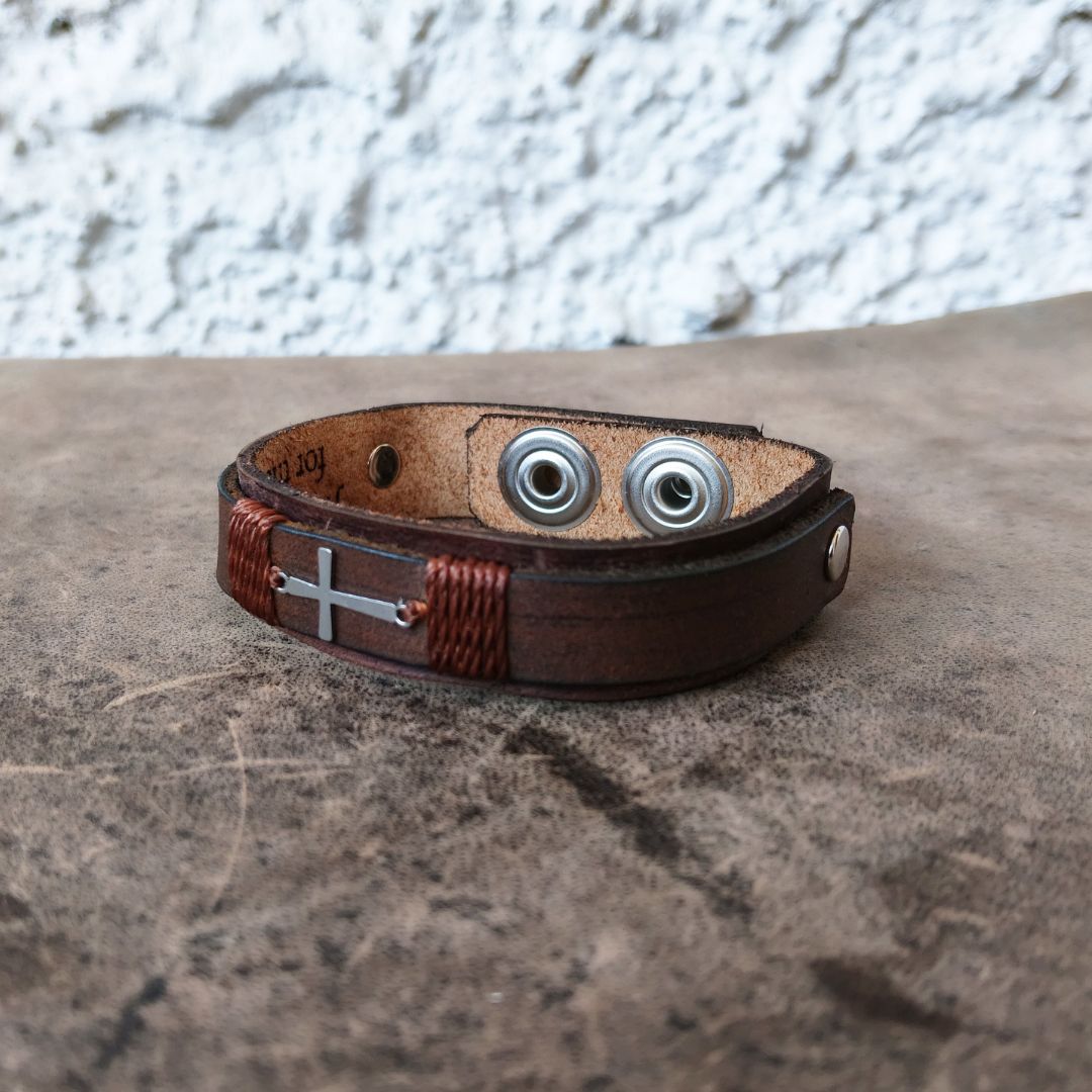 A brown leather wrap bracelet with a silver-toned cross charm and two silver-toned metal eyelets, resting on a textured gray stone surface.