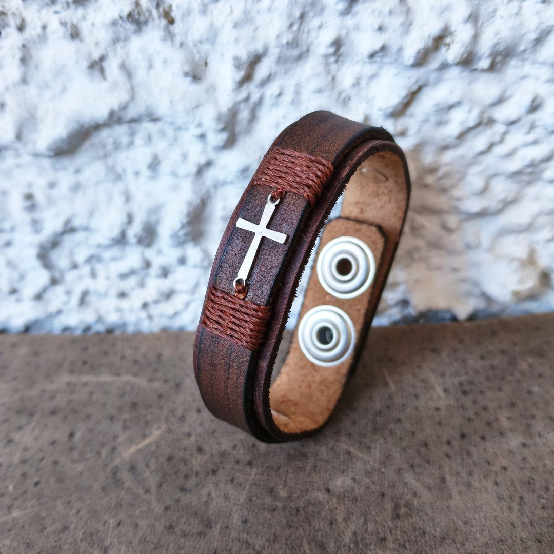 A brown leather wrap bracelet with a silver-toned cross charm, wrapped with brown cord, and featuring two silver-toned metal eyelets on the back, displayed on a textured brown surface against a white stucco wall.