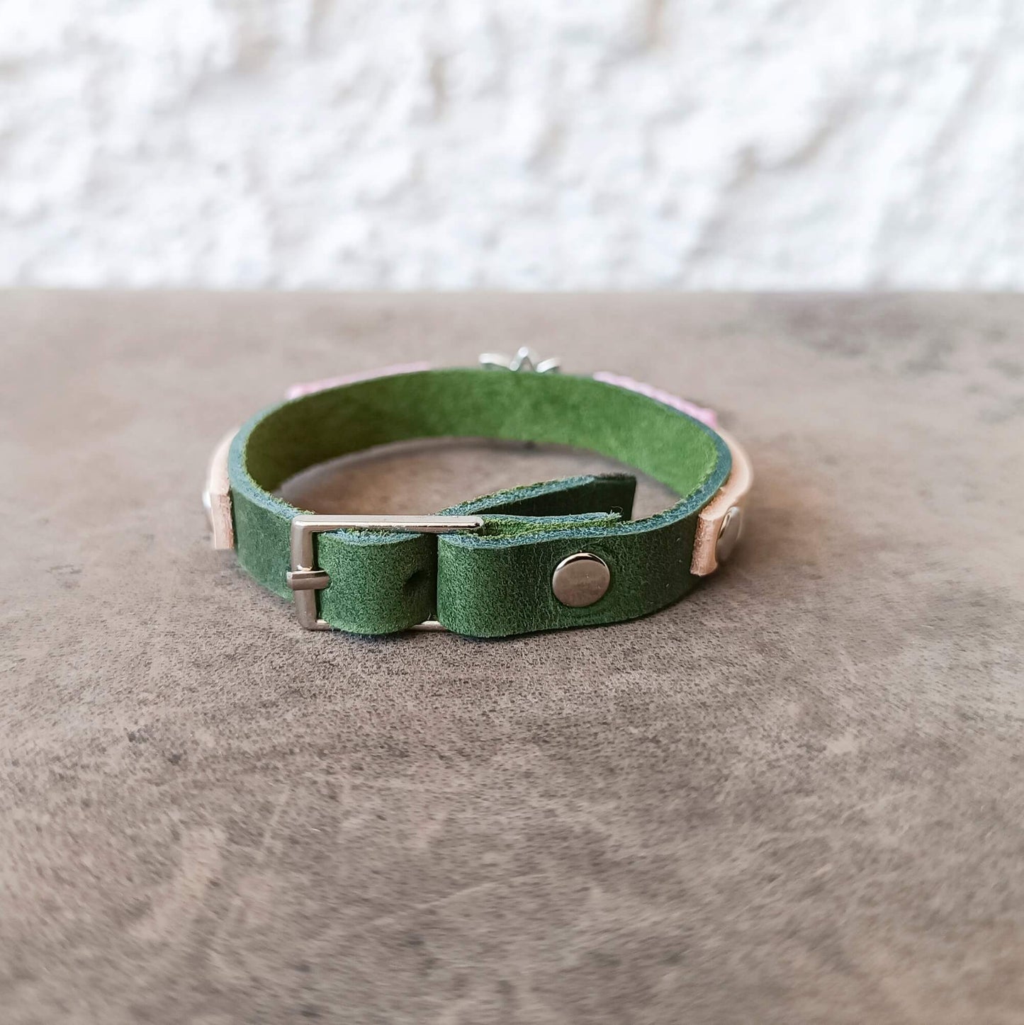 A green leather cuff bracelet with a silver-tone metal snap button and buckle, resting on a textured gray surface.