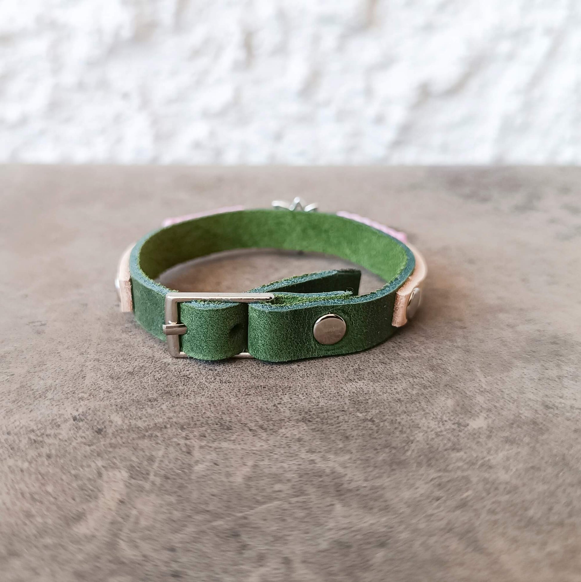 A green leather cuff bracelet with a silver-tone metal snap button and buckle, resting on a textured gray surface.
