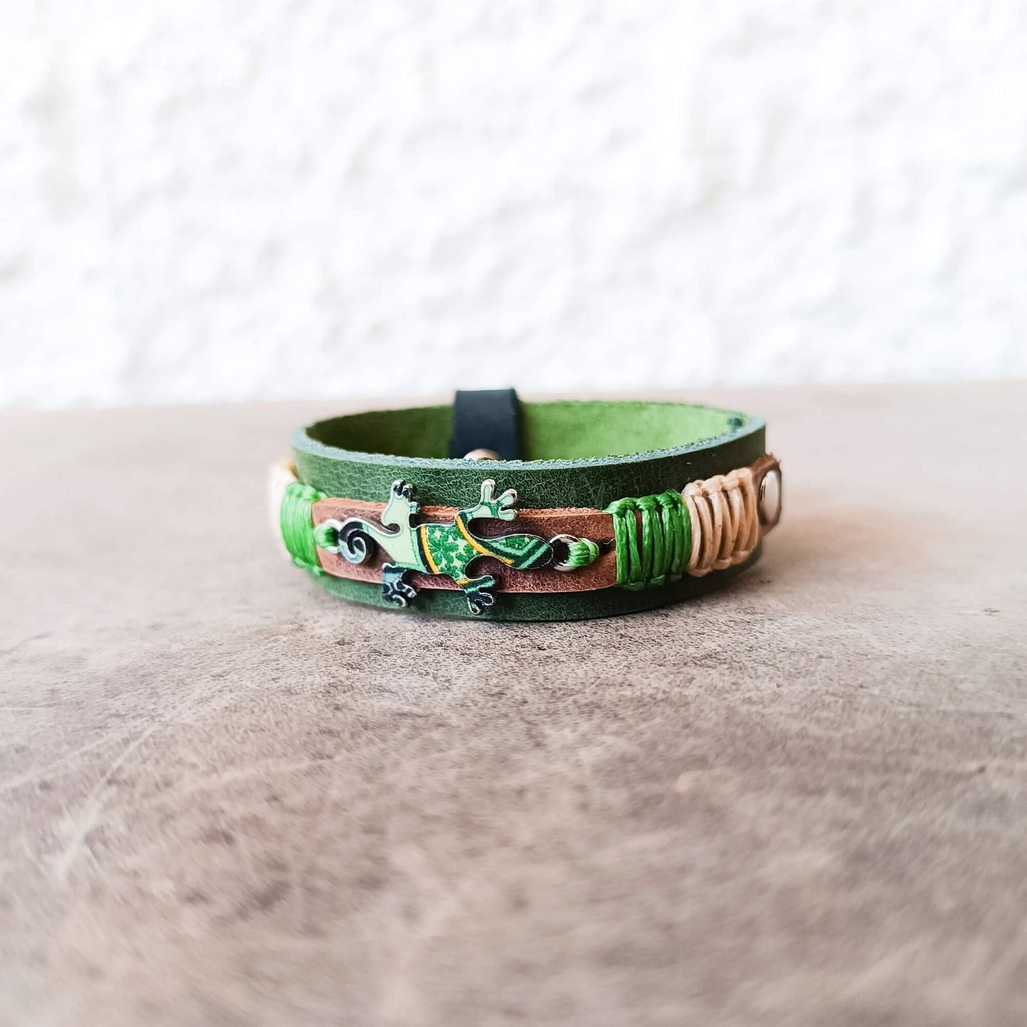 A handmade green leather cuff bracelet featuring a central green gecko charm, a brown leather inlay, and wrapped beige cord accents, resting on a textured brown surface.