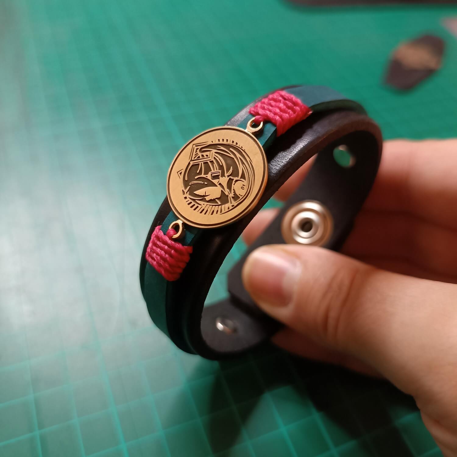 Bracelet with blue strap, pink accents, and gold Hatsune Miku pendant on a green cutting mat - making of