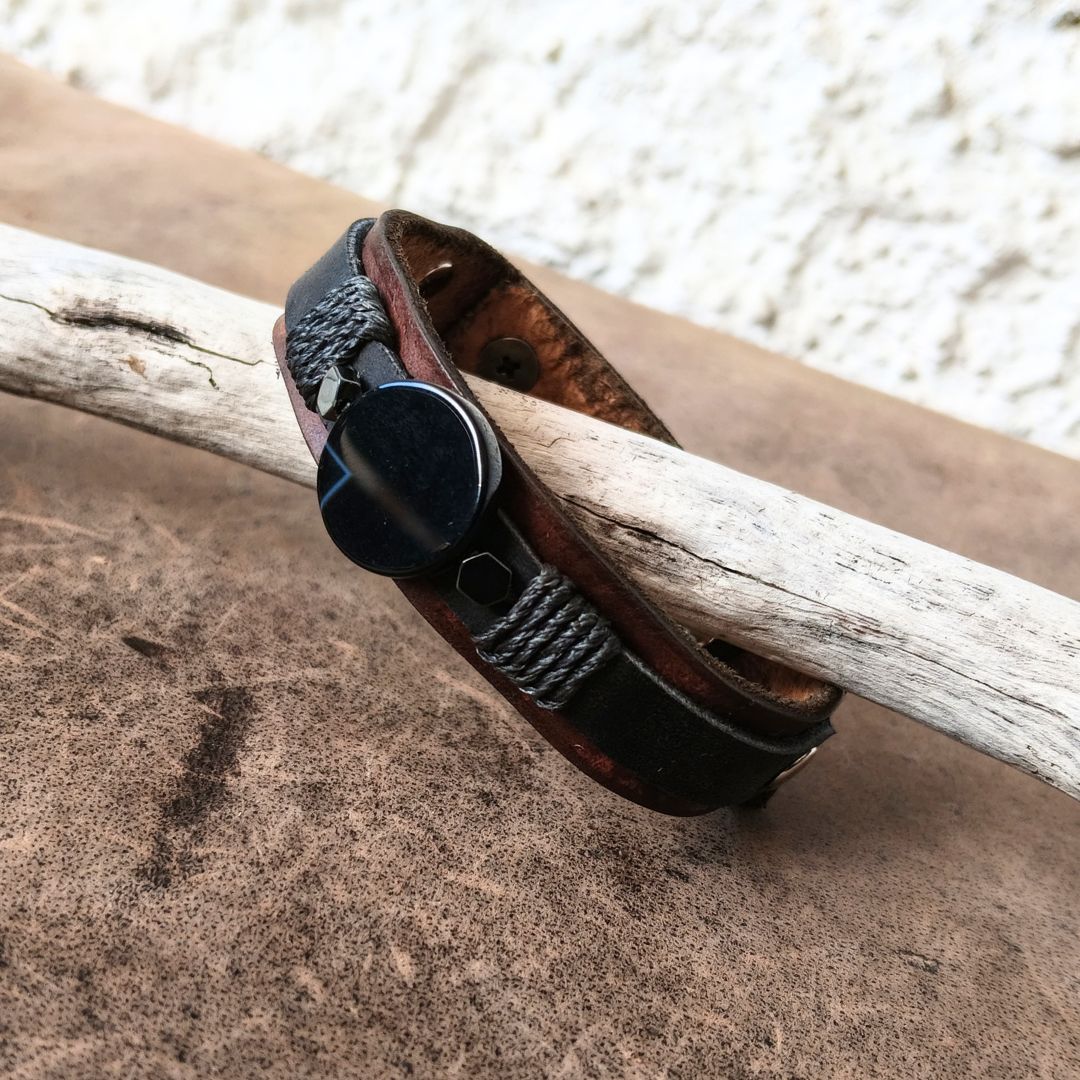 A dark brown leather cuff bracelet with a black agate central piece and braided black cord details, resting on a light-colored driftwood log over a brown textured surface.