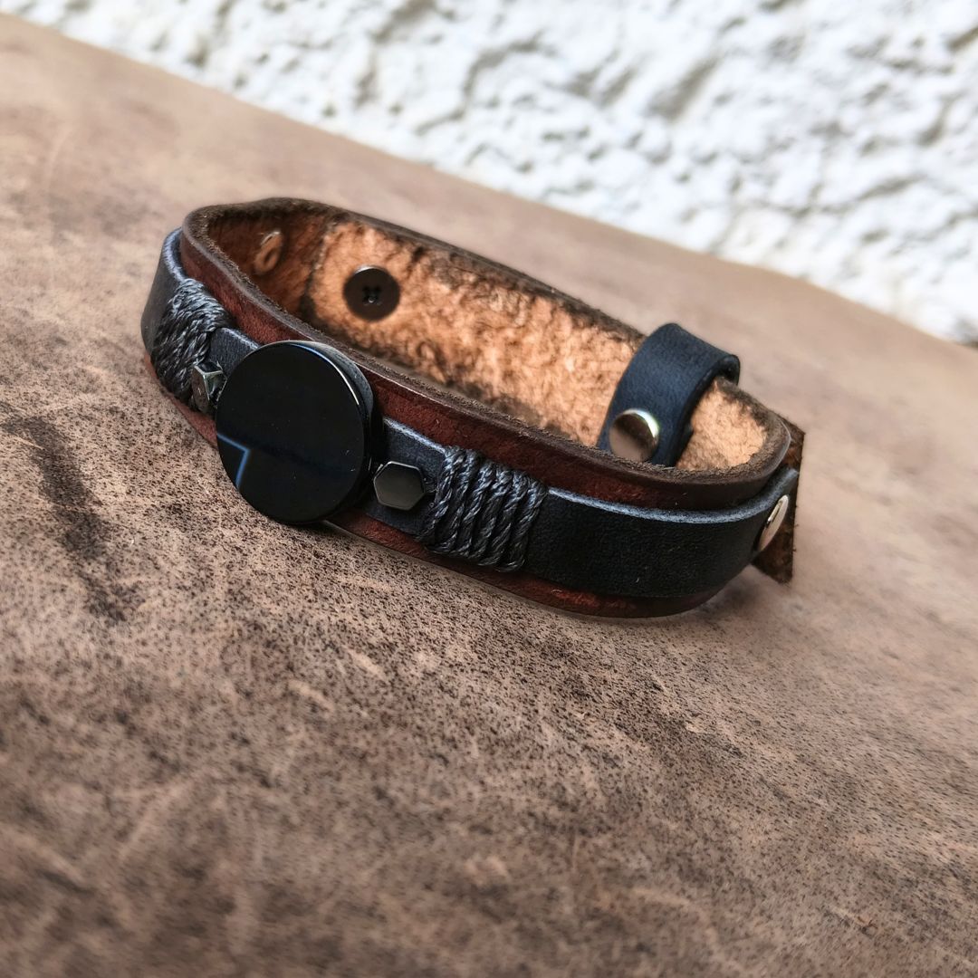 A dark brown leather bracelet with black braided cord detailing, a round black agate charm, resting on a textured brown leather surface.
