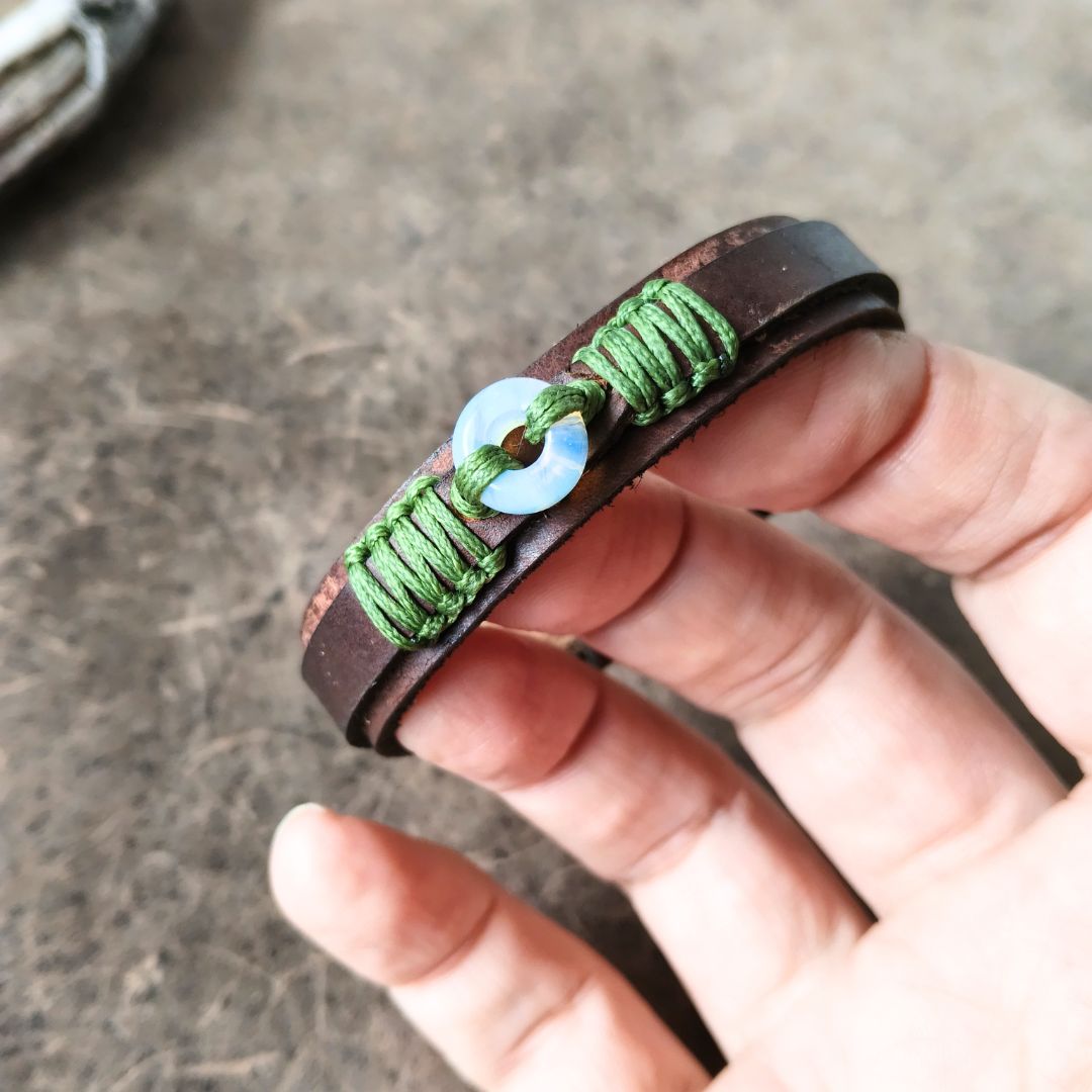 A close-up of a hand holding a brown leather wrap bracelet with green braided cord details and a central moonstone donut bead.