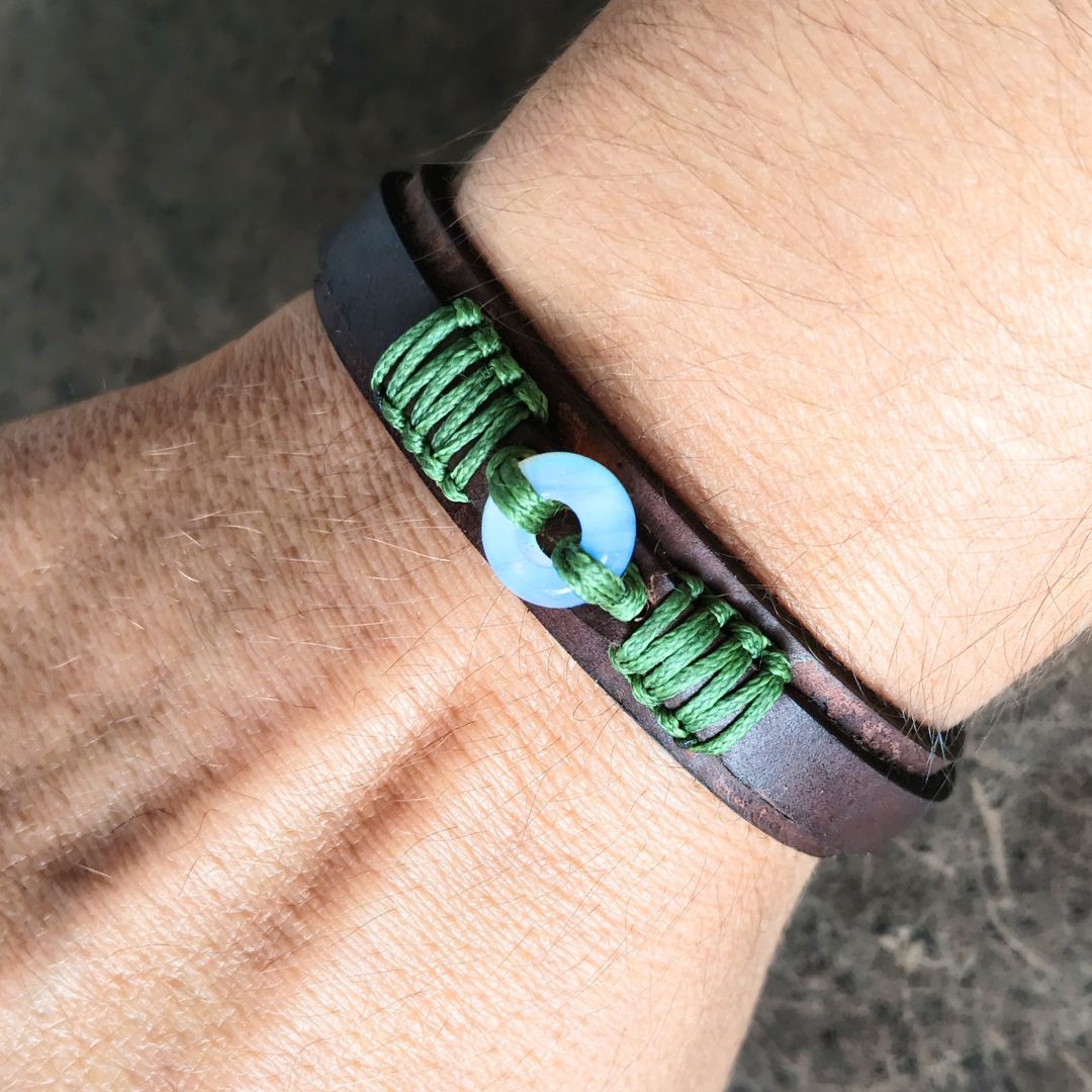 A close-up of a wrist wearing a dark brown leather bracelet with green braided cord accents and a moonstone donut bead.
