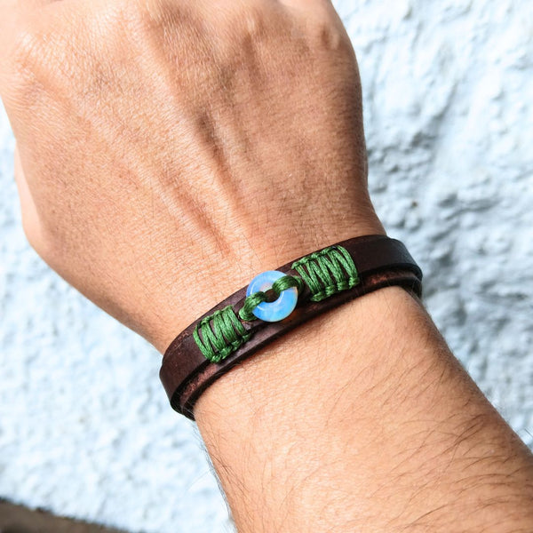 A close-up of a person's wrist wearing a dark brown leather wrap bracelet with green braided cord details and a central Moonstone donut bead.