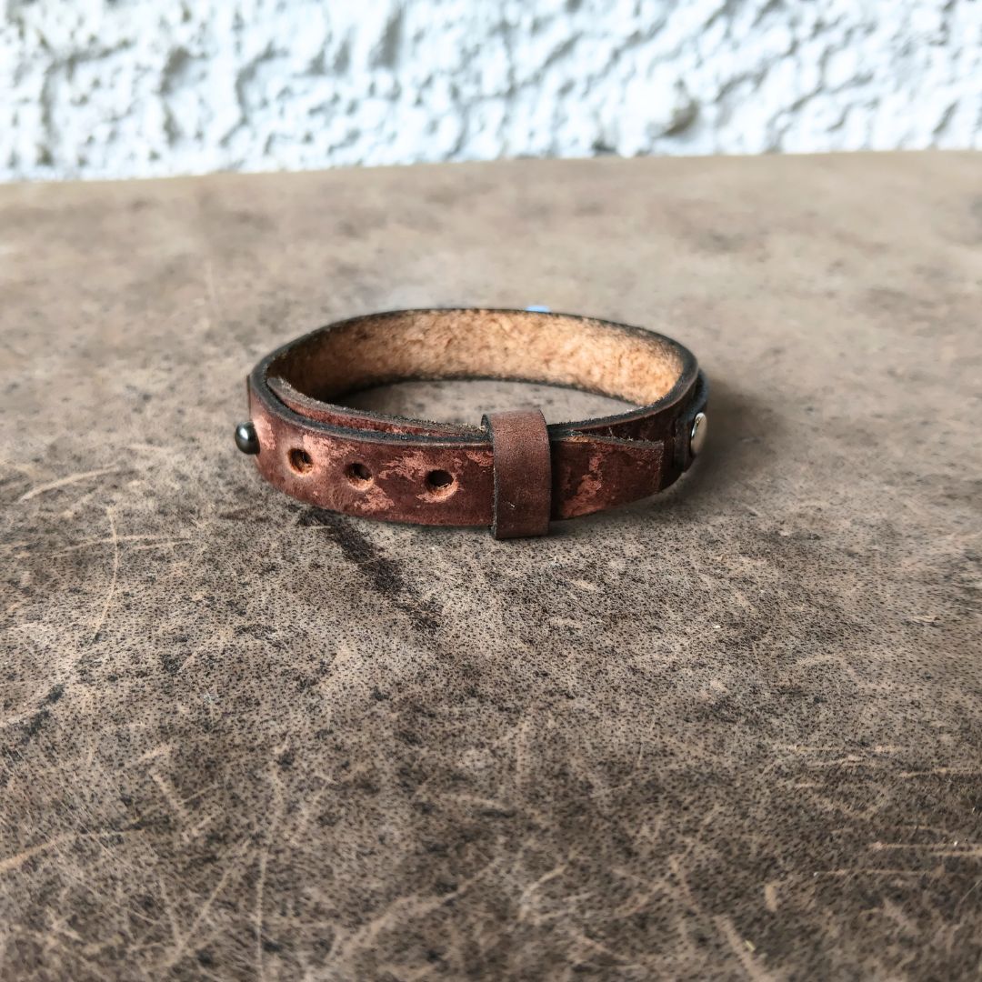 A distressed brown leather cuff bracelet with silver-toned metal rivets and a toggle strap, resting on a textured brown surface.