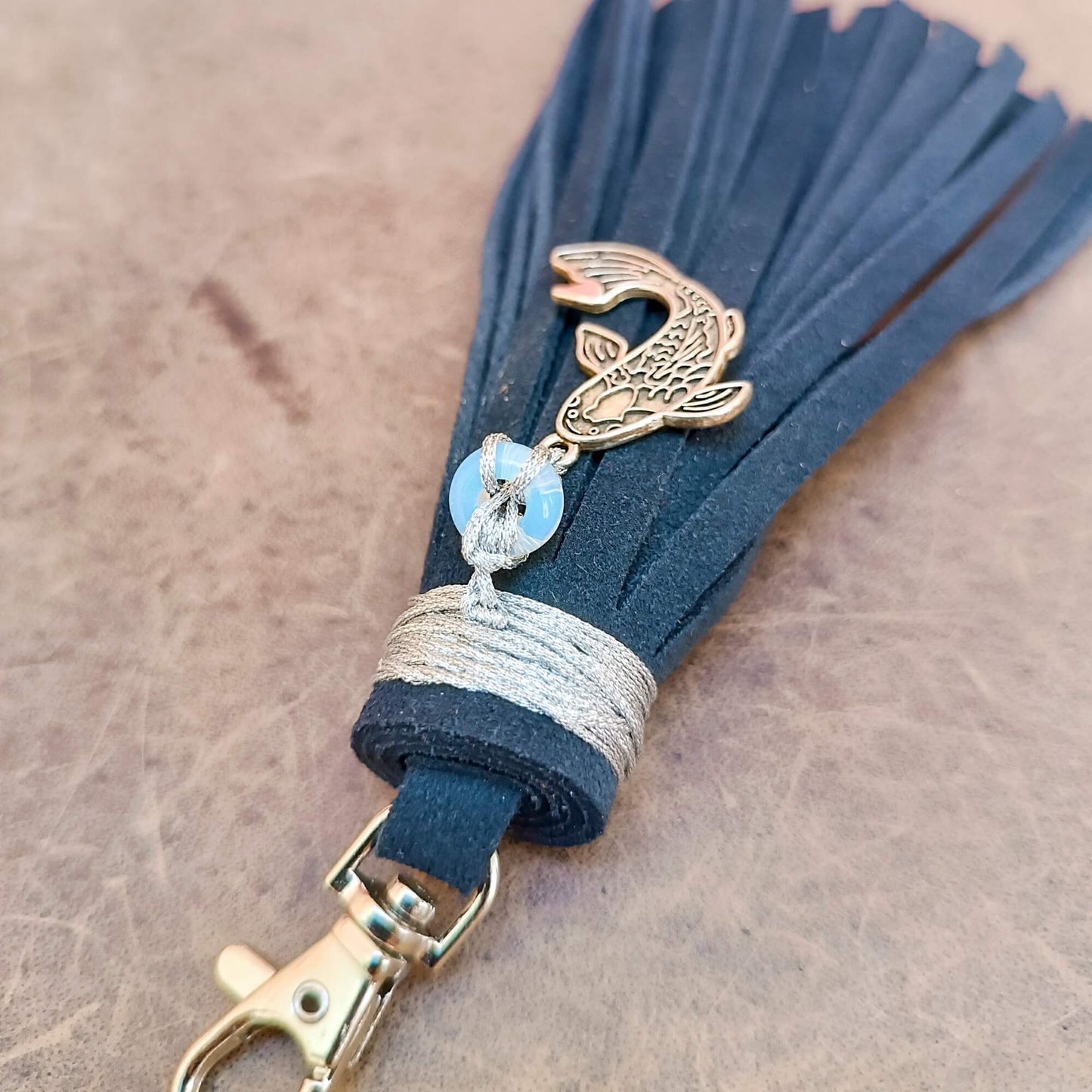A dark blue suede tassel keychain featuring a koi fish charm, a lmoonstone donut bead with a white knot, and a silver wrapped cord, attached to a silver clasp on a textured brown surface.