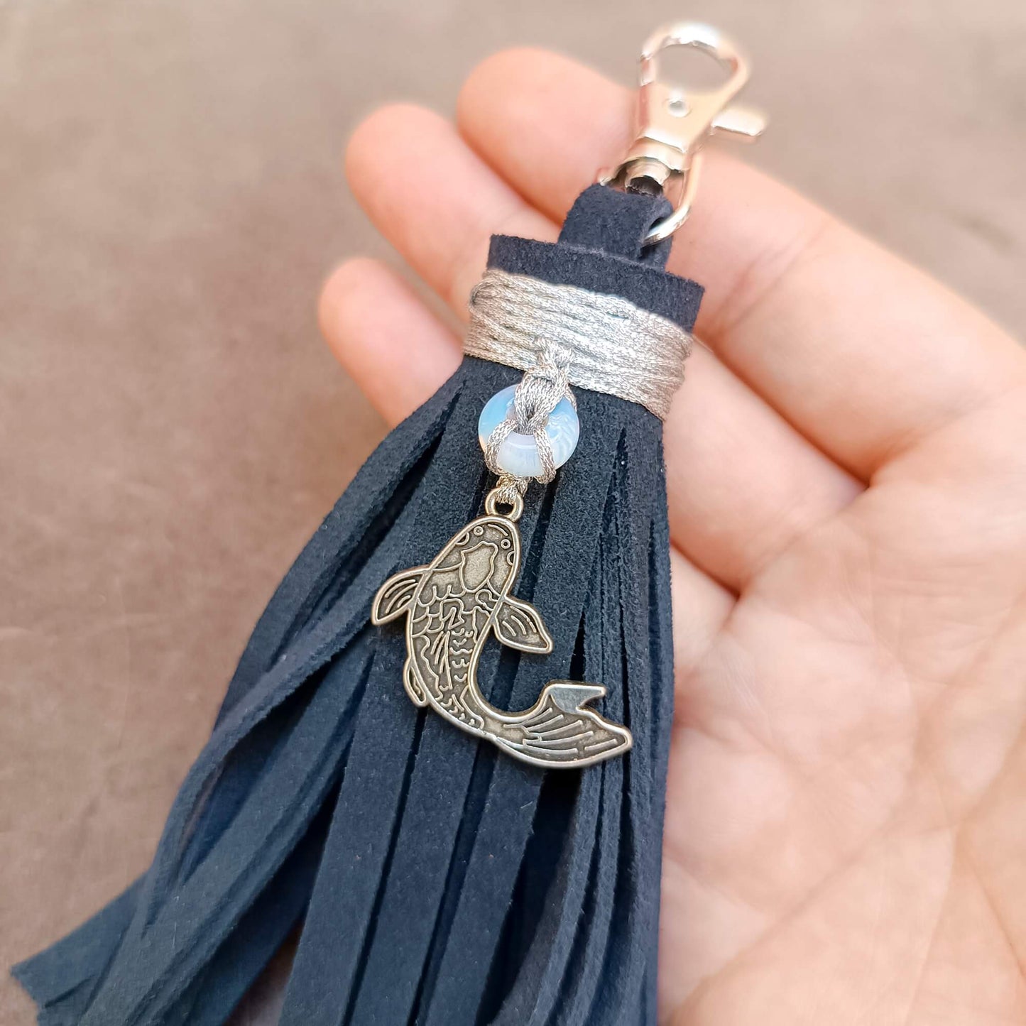 A close-up of a hand holding a dark blue suede tassel keychain with a silver fish charm, a moonstone donut bead, and a silver clasp.