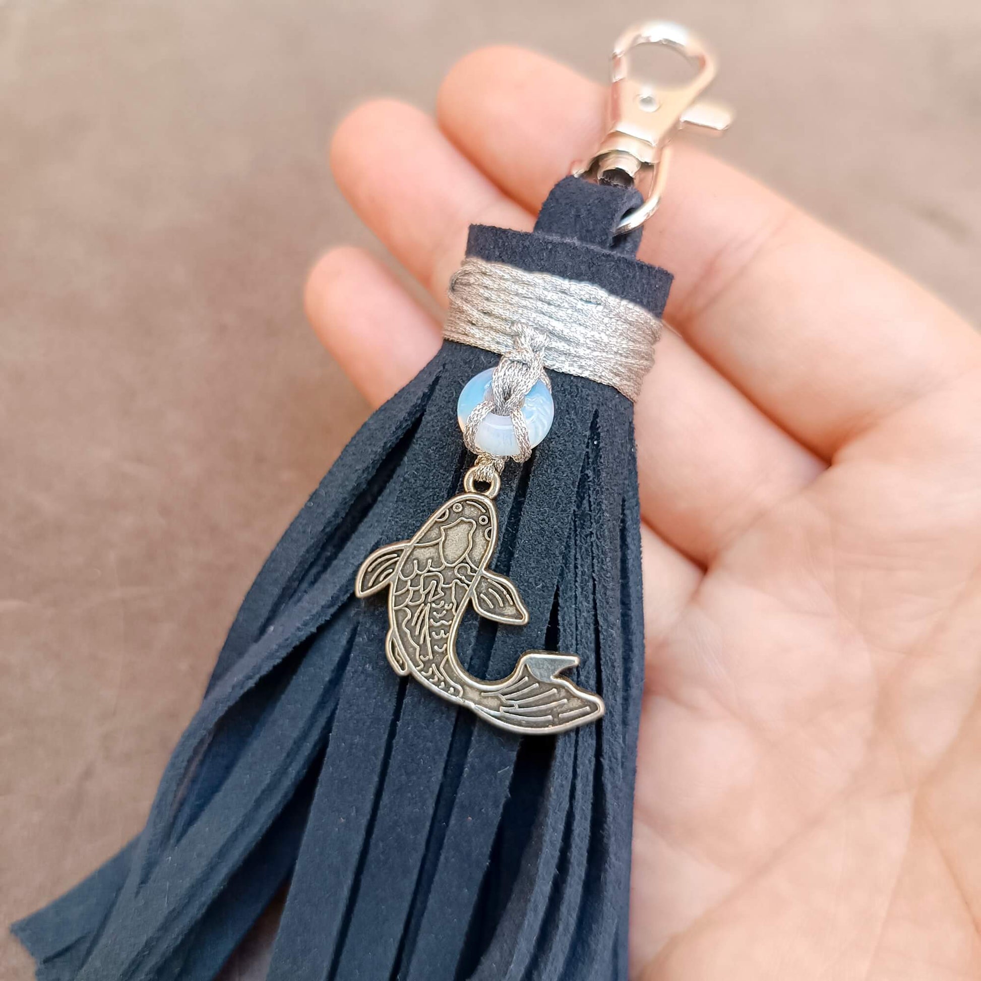 A close-up of a hand holding a dark blue suede tassel keychain with a silver fish charm, a moonstone donut bead, and a silver clasp.