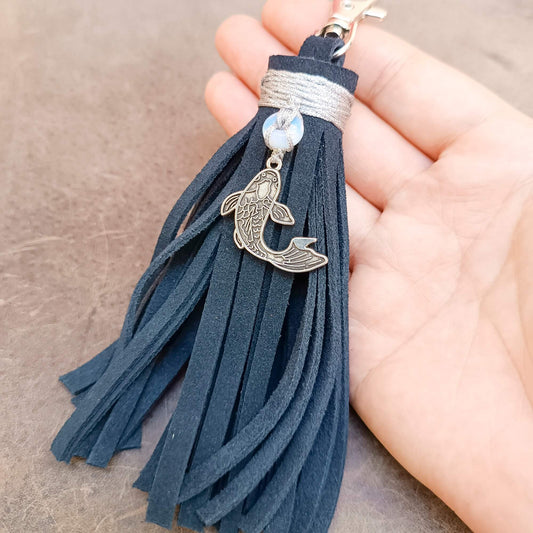 A dark blue suede tassel keychain with a silver fish charm and a small white bead, held in a hand against a neutral background.