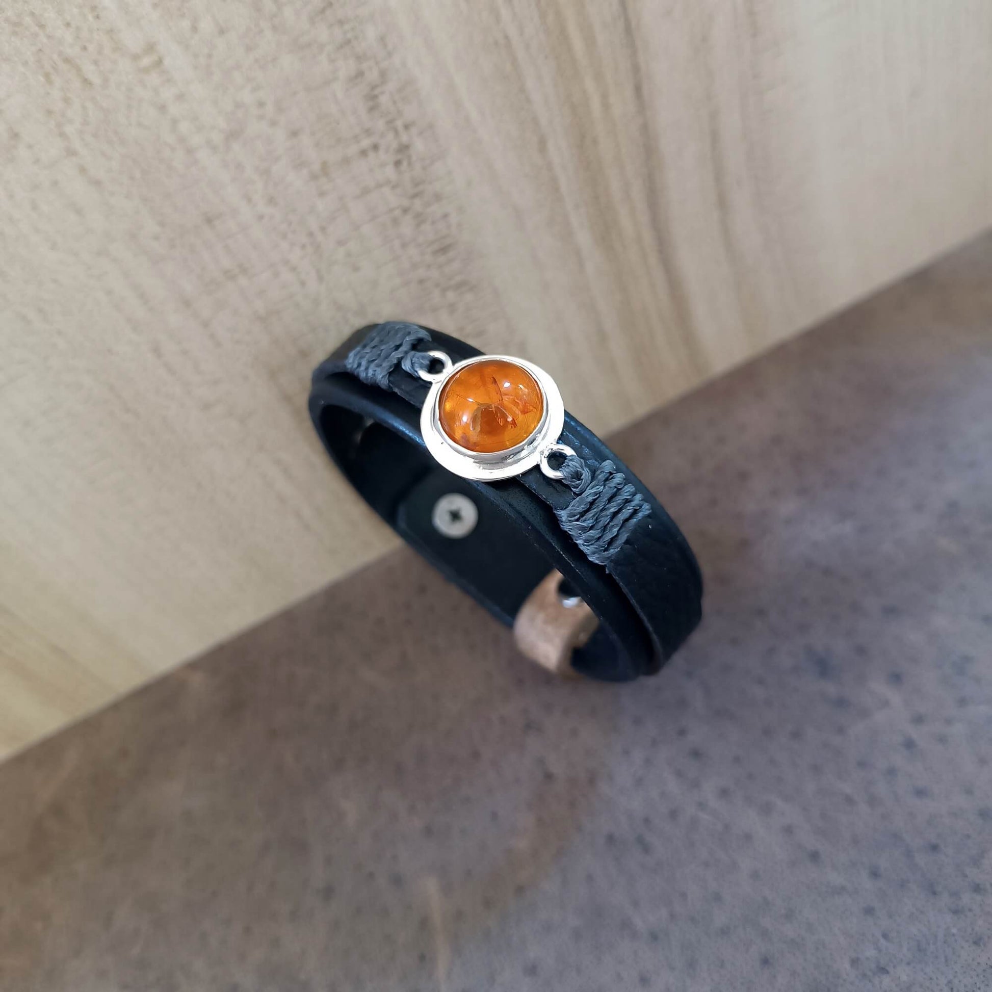 Black leather bracelet with a silver charm featuring an amber stone on a wooden surface.