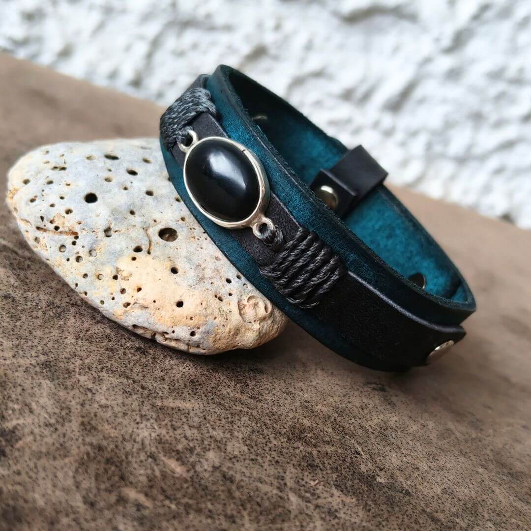 A dark teal and black leather bracelet with a large oval black stone and silver-toned metal accents, styled on a textured brown rock with a small white stone against a white stucco wall background.