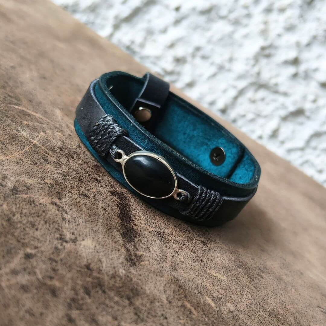 A black leather wrap bracelet with a central oval black stone and silver-toned metal accents, resting on a textured brown surface with a white stucco wall in the background.
