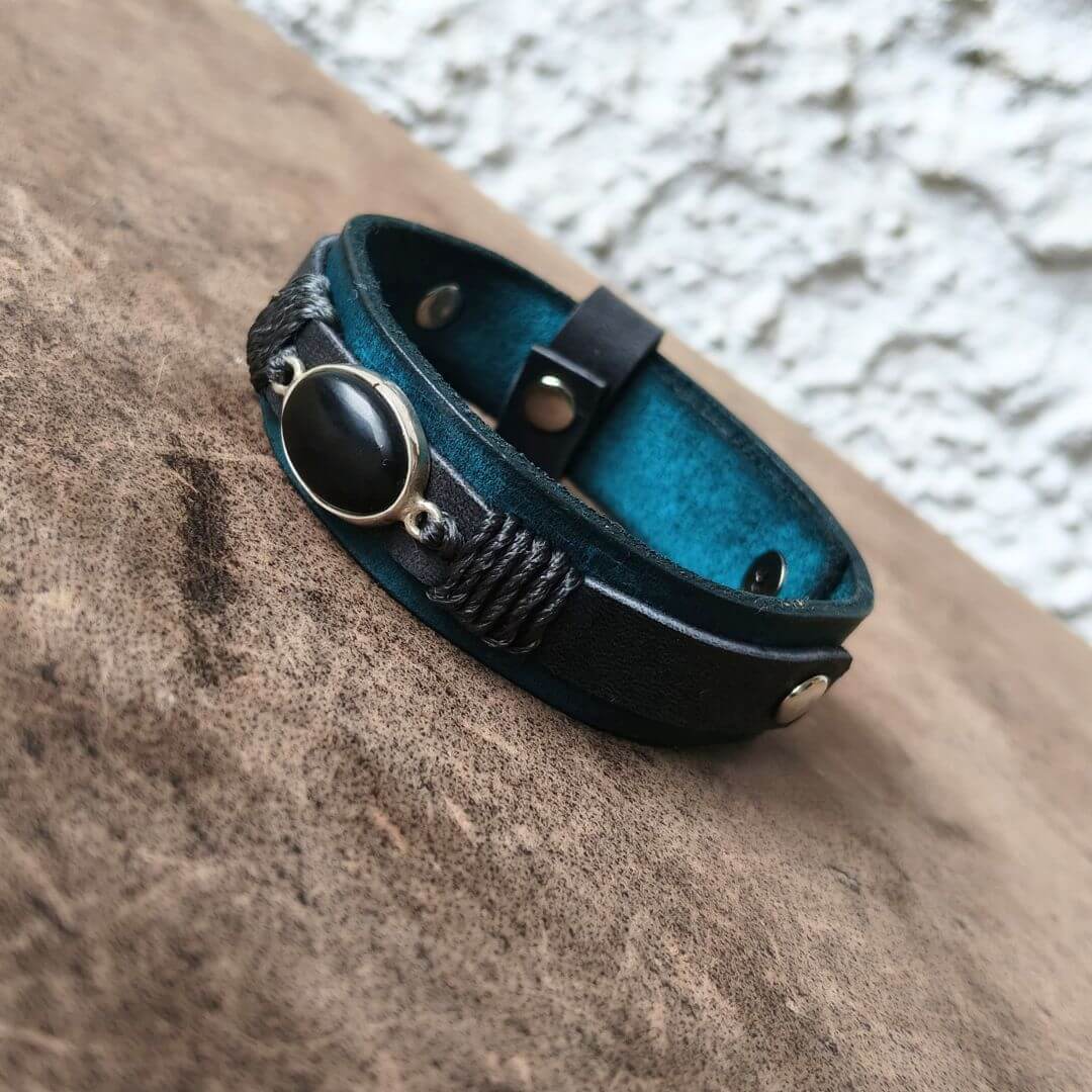 A dark teal leather bracelet with a black oval stone and silver-toned metal accents, resting on a textured brown surface.