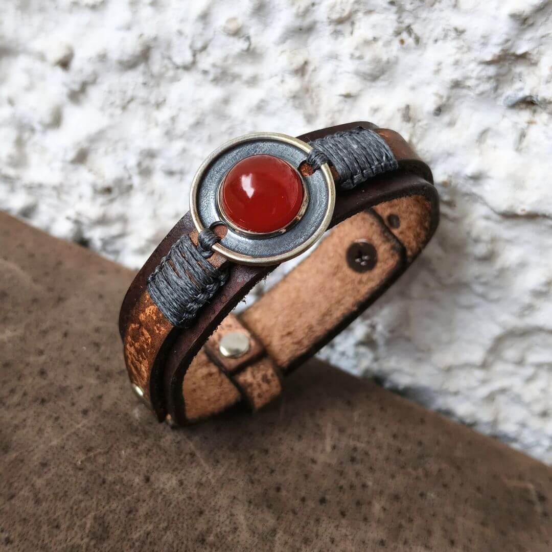 A handmade leather wrap bracelet with a central round red cabochon stone and a textured silver metal bezel, resting on a brown leather surface against a white stucco wall.