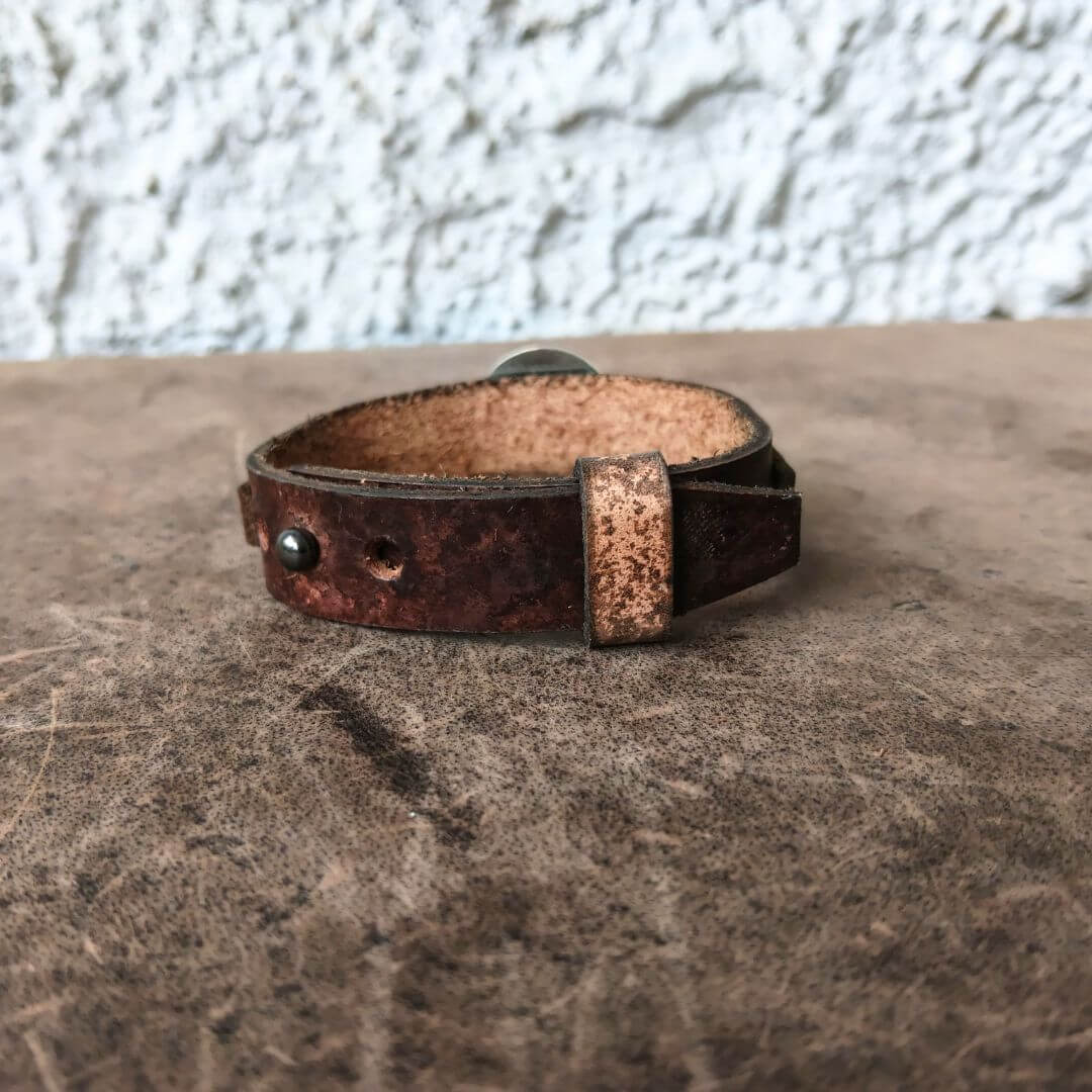 A rustic brown leather cuff bracelet with a distressed finish, a textured rectangular metal accent, and a silver-toned snap closure, resting on a textured brown surface against a white stucco wall.