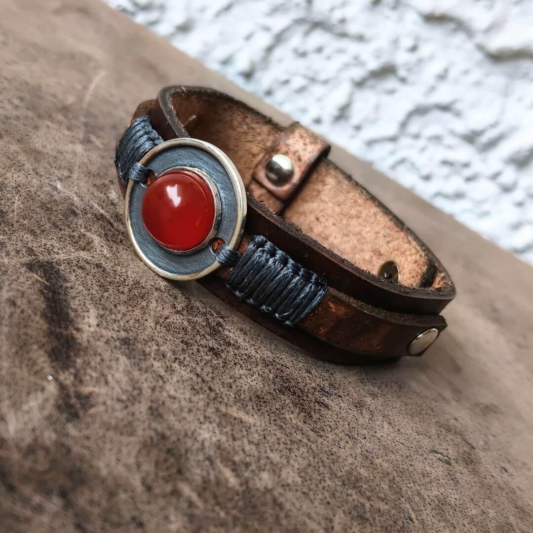 A handmade leather wrap bracelet with a central round bezel containing a red cabochon stone, accented by blue braided cord and brass hardware, resting on a textured brown surface.