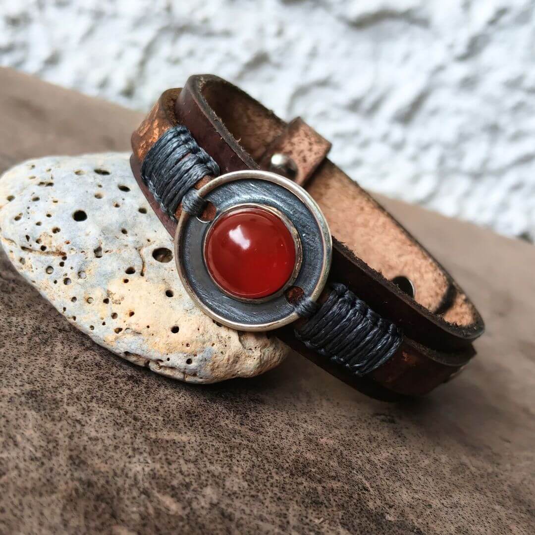 A handmade leather bracelet with dark brown wrap, a silver metal ring, and a round red carnelian stone centerpiece, resting on a textured brown surface next to a light-colored stone.