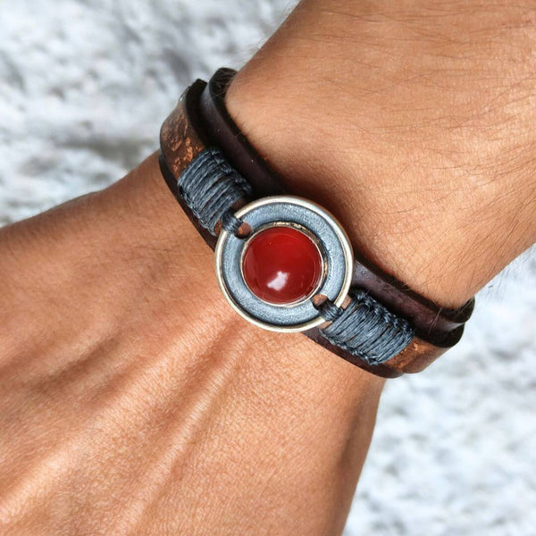 A close-up of a person's wrist wearing a dark brown leather wrap bracelet with a circular silver centerpiece featuring a polished red stone.