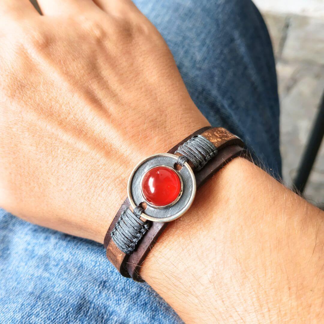 A close-up of a person's wrist wearing a dark brown leather wrap bracelet with a circular bezel holding a round red cabochon stone.