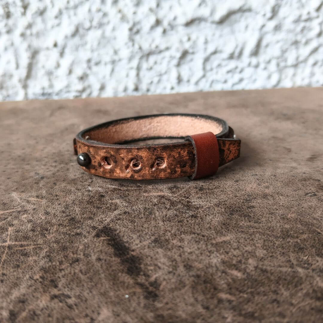 A rustic brown leather cuff bracelet with a textured, distressed finish, featuring a small metal rivet and a contrasting reddish-brown leather wrap near the closure, displayed on a textured brown surface.