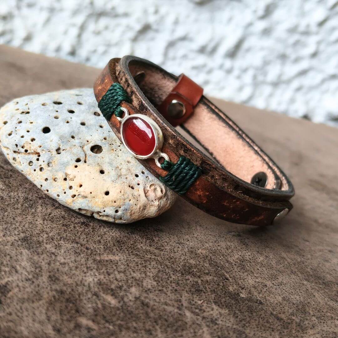 A handmade leather cuff bracelet with a polished red cabochon stone and green cord accents, resting on a textured stone surface.