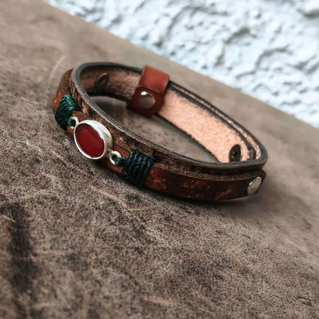 A handmade leather bracelet with a distressed brown finish, featuring a central oval red stone in a silver bezel, accented with green cord wraps and silver studs, resting on a textured brown stone surface.