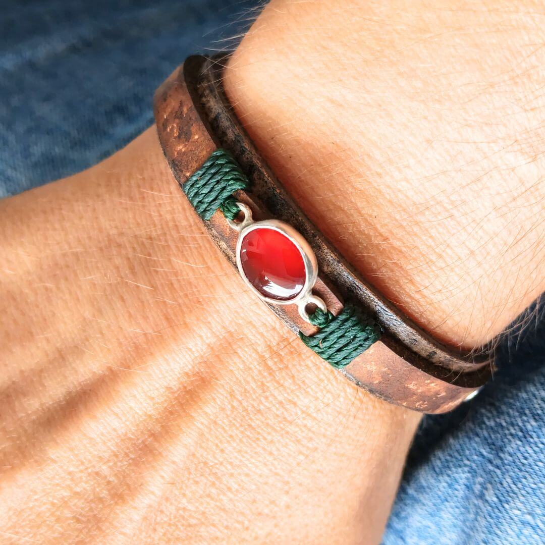 A close-up of a wrist wearing a handmade bracelet with a dark brown leather wrap, green braided cord accents, and a central oval red stone in a silver-toned bezel.
