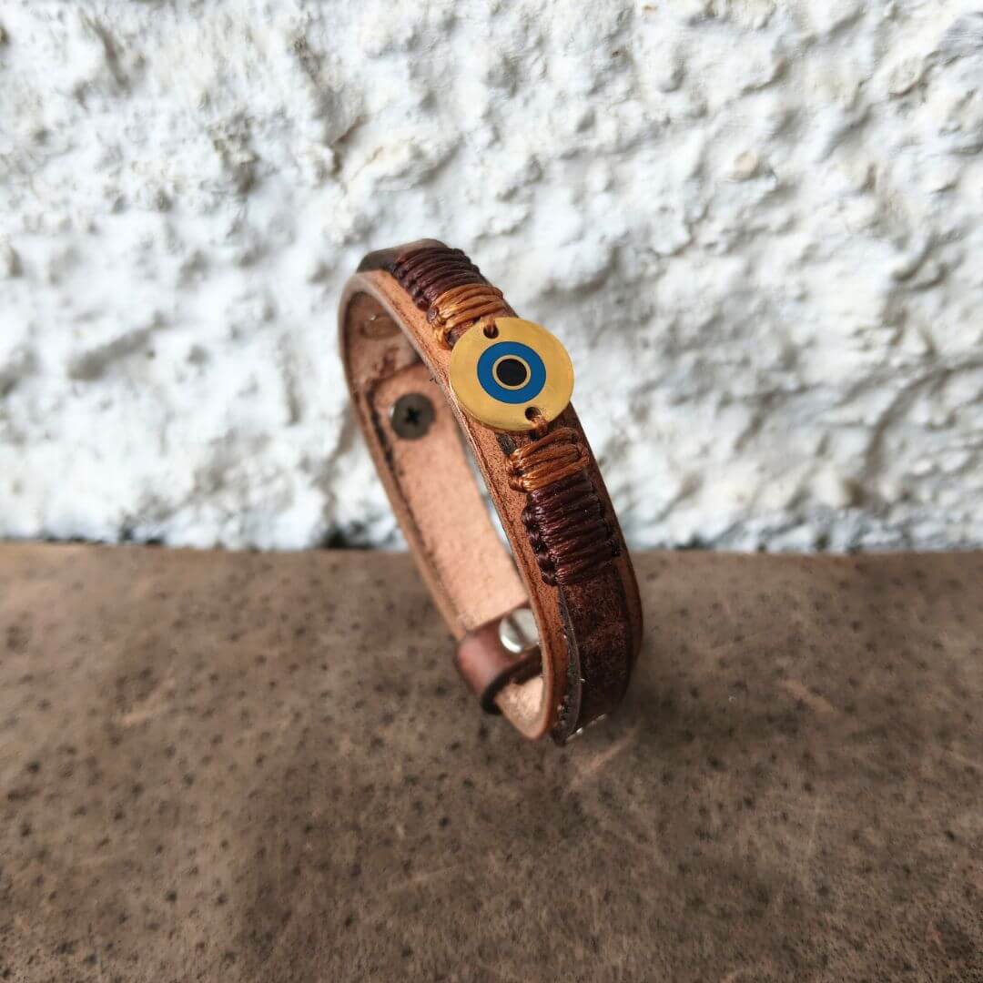 A brown leather wrap bracelet with a textured woven band and a round evil eye charm, displayed on a textured brown surface against a white stucco wall.