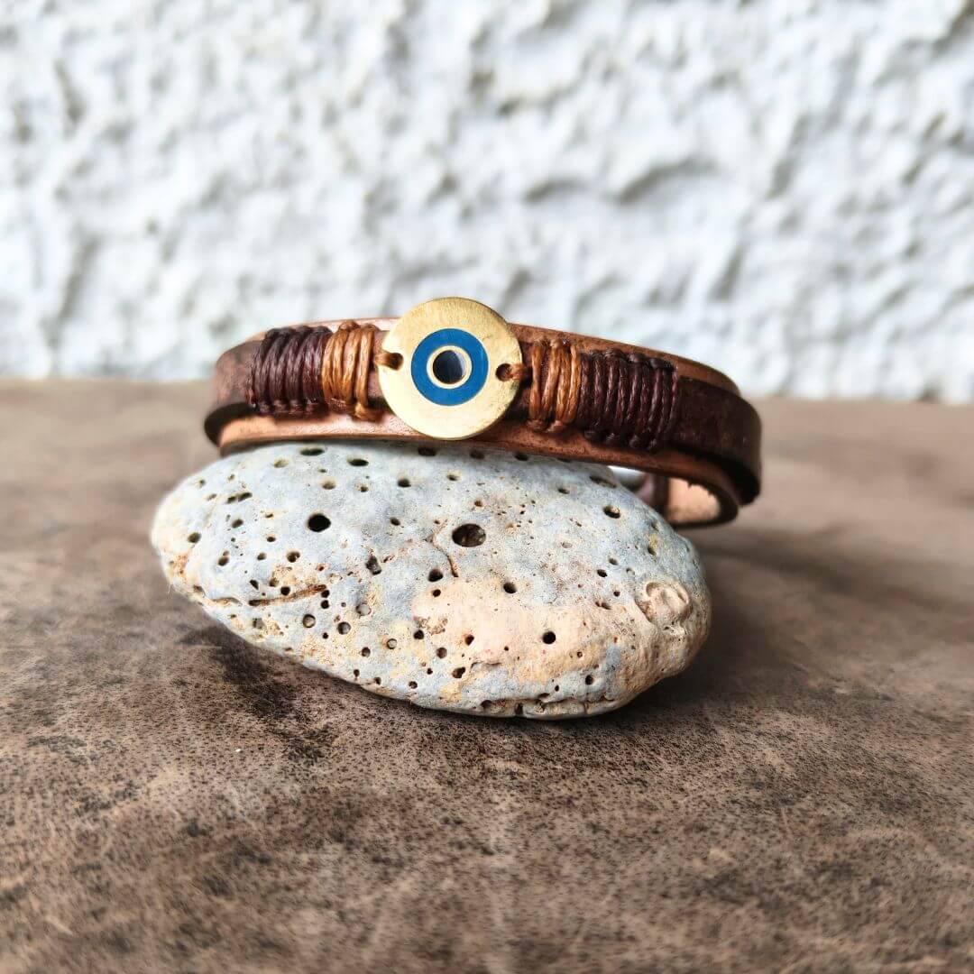 A handmade leather cuff bracelet featuring a circular evil eye charm in blue, and gold. The bracelet is displayed on a small speckled stone against a textured gray background.