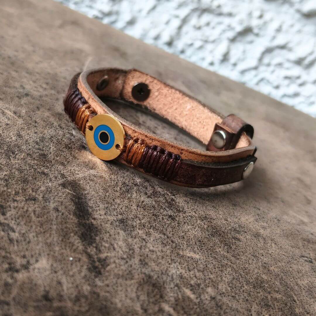 A brown leather wrap bracelet with a circular gold-toned evil eye charm featuring blue and black details, resting on a textured gray stone surface.