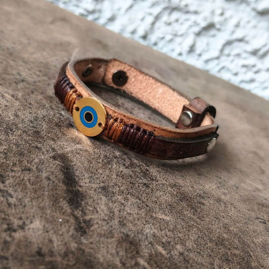 A brown leather wrap bracelet with a circular gold-toned evil eye charm featuring blue and black details, resting on a textured gray stone surface.