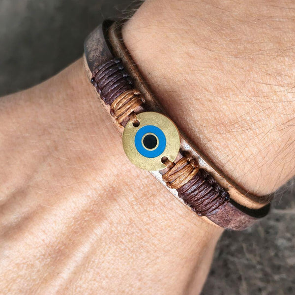 A close-up of a person's wrist wearing a dark brown leather wrap bracelet with a round gold evil eye charm 