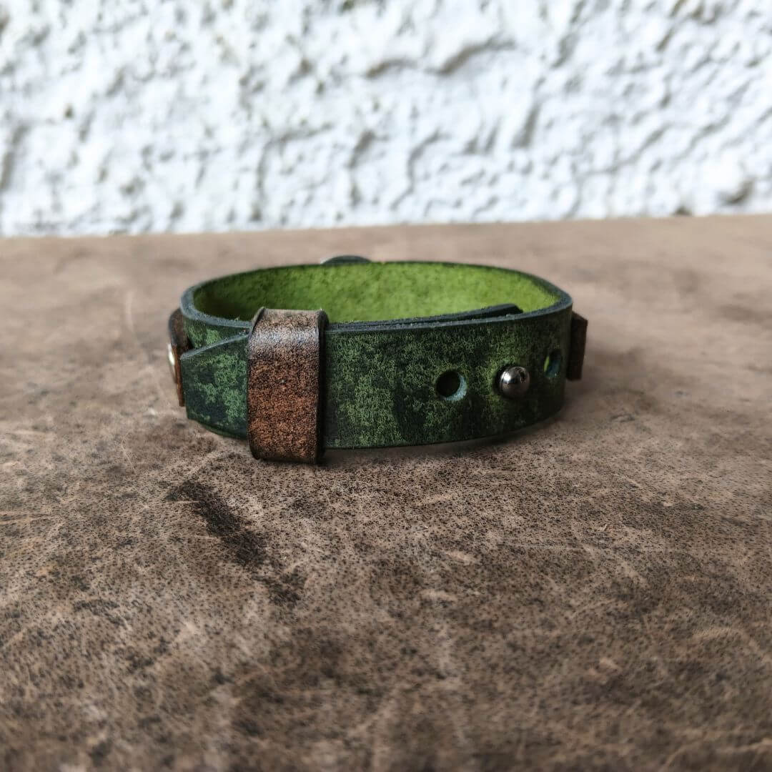 A handmade leather cuff bracelet with a distressed green and black exterior, a brown leather tab, and a bright green interior, resting on a textured brown surface.