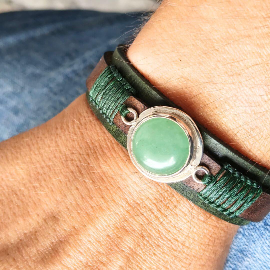 A close-up of a person's wrist wearing a dark green leather wrap bracelet with a central round green stone set in a silver-toned bezel.