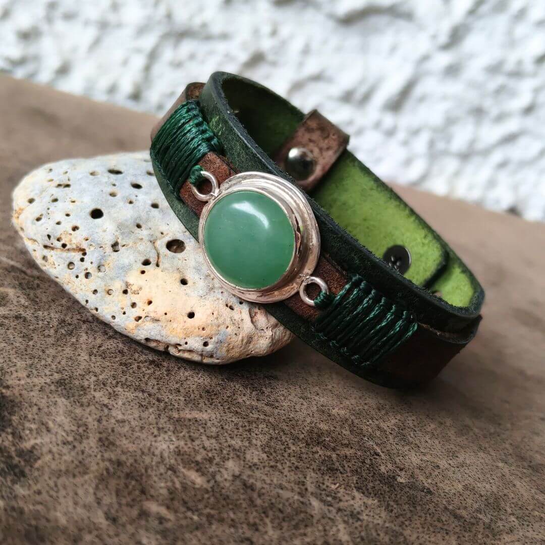 A handmade wrap bracelet made of dark brown leather with green braided cord accents and a large oval green stone in a silver-toned bezel setting, resting on a textured brown surface next to a white stone.