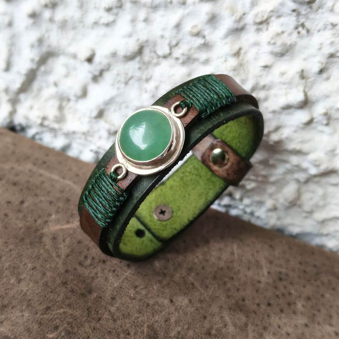 A green leather wrap bracelet with a central oval green stone set in a silver-toned bezel, resting on a brown textured surface against a white stucco wall.