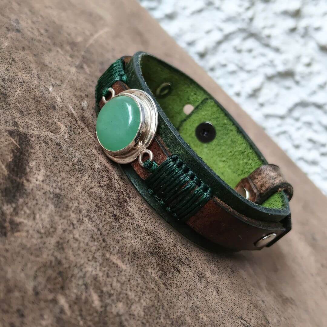 A handmade leather bracelet with a green cabochon stone and dark green braided cord, resting on a textured brown surface.