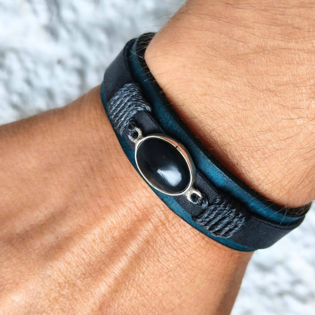 A close-up of a person's wrist wearing a dark teal and black leather wrap bracelet with a silver-toned oval bezel containing a black stone.