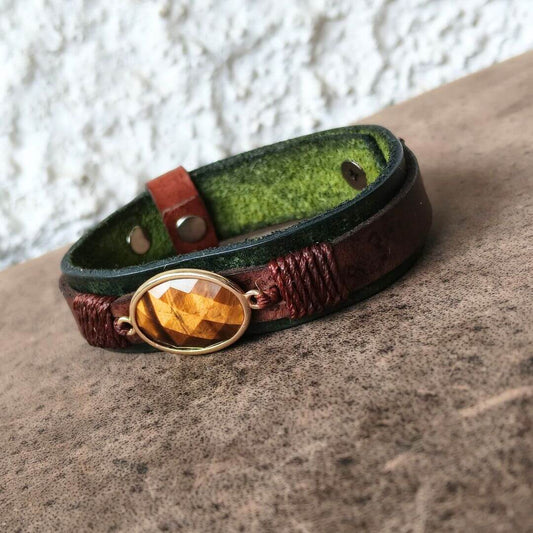 A handmade leather wrap bracelet with a faceted tiger's eye gemstone in a gold bezel, resting on a textured brown surface against a white stucco wall.