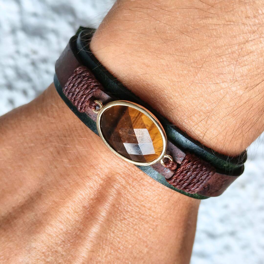 A close-up of a person's wrist wearing a multi-layered leather bracelet. The bracelet features a dark brown leather band, a black leather band, and a central oval bezel containing a tiger's eye stone.