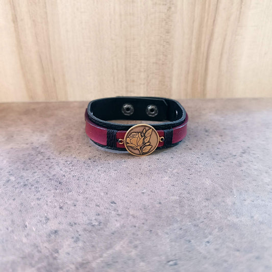 Black and red leather bracelet with a gold brass rose pendant on a gray surface.