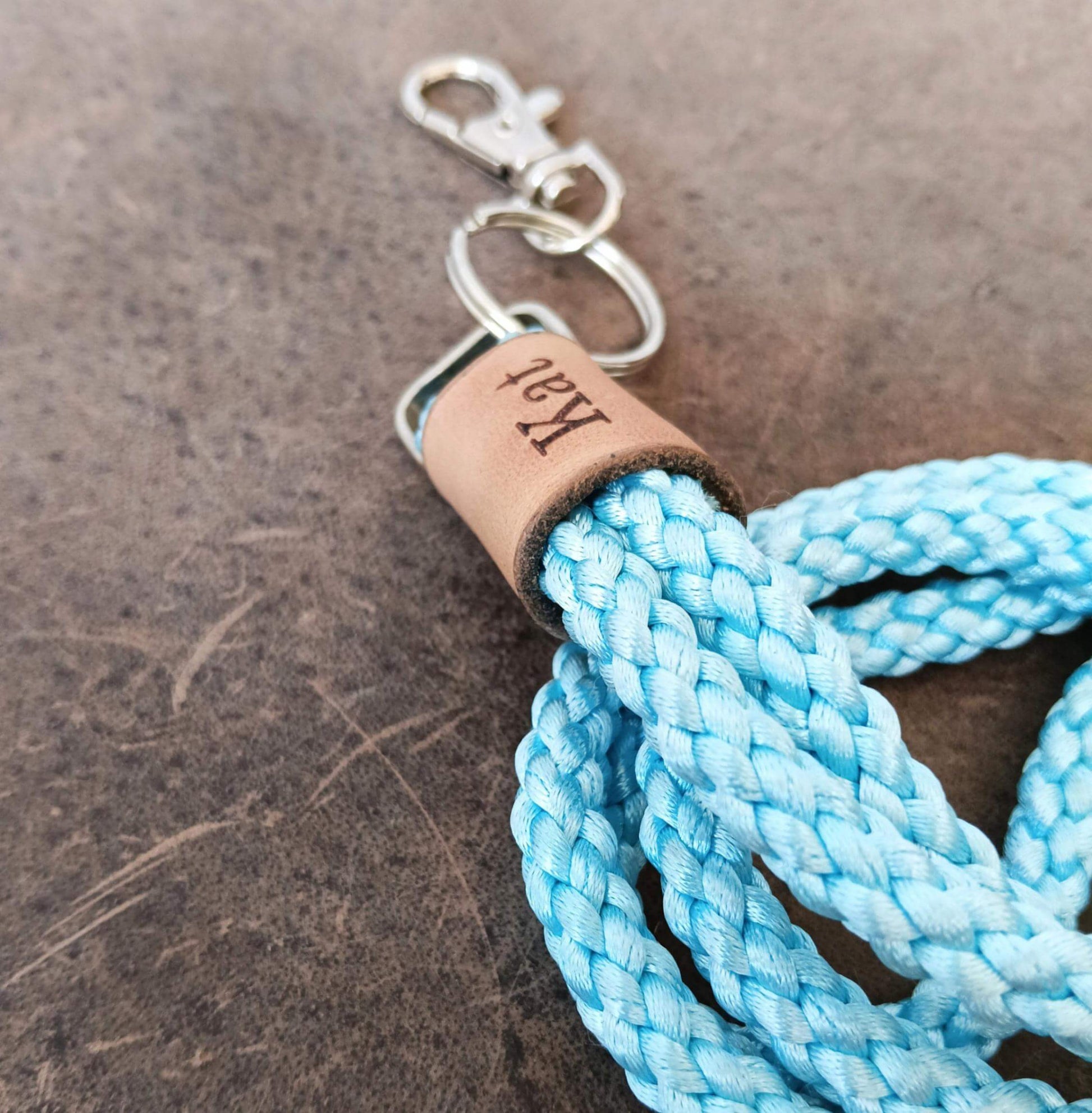 A close-up of a light blue braided rope lanyard with a silver metal clasp and a tan leather wrap engraved with the name 'Kai', set against a dark textured background.