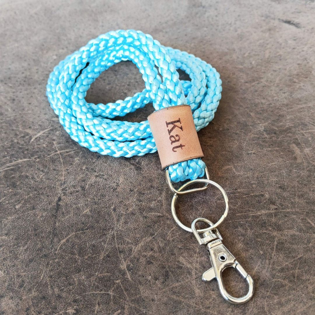 A light blue and white braided rope lanyard with a brown leather tag engraved with the name 'Kat' and silver metal key rings and clasp, displayed on a dark textured surface.